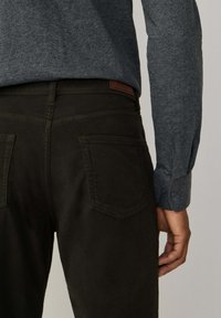 Pantalones verde oscuro con una textura suave, que cuentan con dos bolsillos traseros y una etiqueta marrón en la cintura, combinados con una camisa gris de manga larga.