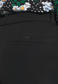 Pantalons noirs ajustés avec une texture lisse, comportant une poche arrière et une petite étiquette métallique attachée pour le branding.