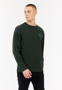 Donkergroen katoenen t-shirt met lange mouwen en een ronde hals. Heeft een klein, lichtgroen logo op de linkerborst. Sluit strak aan.