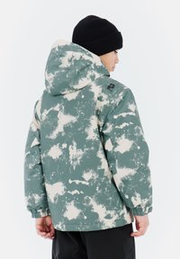 Groene en crèmekleurige jas met een spetterpatroon en capuchon, gemaakt van waterafstotend materiaal; beschikt over elastische manchetten en een logo op de schouder.