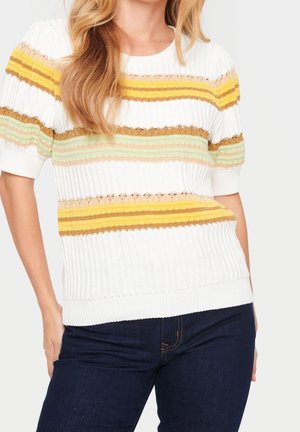 Kobieta w białym dzianinowym swetrze z krótkim rękawem, ozdobionym żółtymi, brązowymi i zielonymi poziomymi paskami, do którego dobrała ciemnoniebieskie dżinsy.