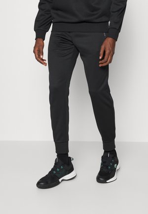 Homme portant un pantalon de jogging noir fuselé, des chaussures de sport noires avec semelles blanches, debout devant un fond clair uni.