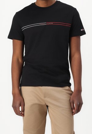 Mężczyzna ubrany w czarną koszulkę Tommy Jeans z białymi i czerwonymi paskami na wysokości klatki piersiowej oraz beżowe spodnie, z rękami swobodnie opuszczonymi wzdłuż ciała.