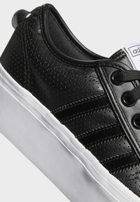 Černé syntetické tenisky s perforovaným svrškem, bílou gumovou podrážkou a černými tkaničkami. Ma mají tři černé pruhy po straně a poutko.