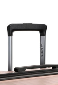 Maniglia del trolley estensibile in metallo argentato con impugnatura in plastica nera su una valigia rosa chiaro, contrassegnata con il marchio "SURI FREY" sullo stelo della maniglia.