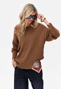 Bruine gebreide oversized trui met geribde zoom en mouwen, gecombineerd met donkere jeans, met een patroonsjaal die uit de zak steekt.