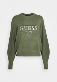 Oliwkowy sweter z miękkiej dzianiny, o szerokim kroju, długich rękawach i trójkątnym logo "GUESS" ozdobionym cyrkoniami.