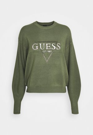 Pull en olive en maille douce, affichant une silhouette ample, des manches longues et un logo "GUESS" triangulaire orné de rhinestones.