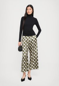 Zwarte turtleneck, met lange mouwen. Hoge taille broek met brede pijpen in groen, crème en zwart. Model houdt een kleine zwarte tas vast en draagt zwarte hakken.
