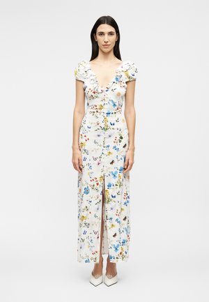 Femme portant une robe maxi blanche à fleurs avec des manches bouffantes, un décolleté en V profond, des boutons à l'avant, une fente frontale et des chaussures blanches pointues.