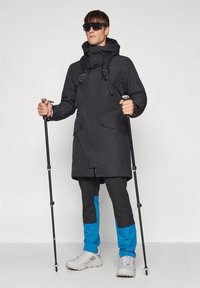 Juoda neperšlampama striukė su gaubtu, kurioje yra dvi kišenės priekyje ir reguliuojamos juostos. Derinama su juodomis ir mėlynomis kelnėmis bei žygių lazdomis.