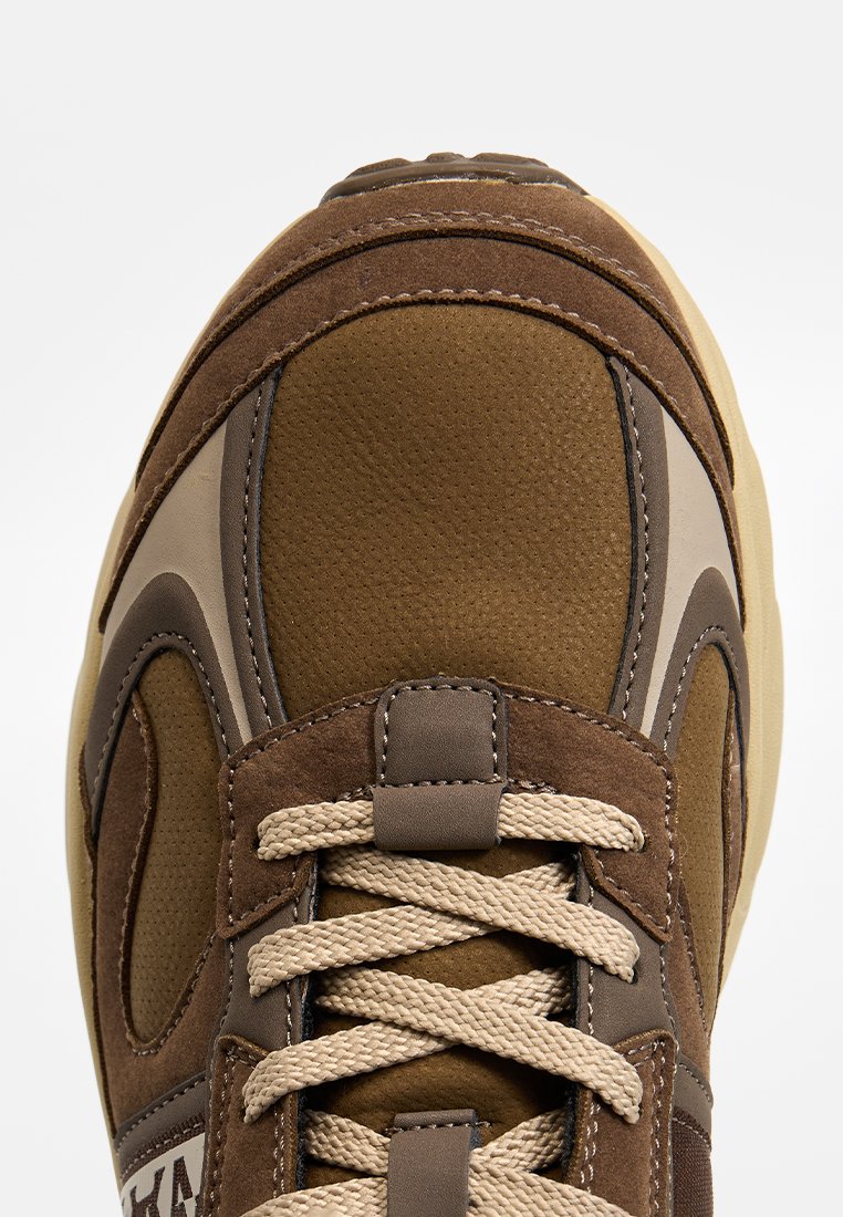 Zapatilla marrón con parte superior texturizada de ante y malla, cordones beige y suela de goma, con detalles de diseño curvados y costuras.