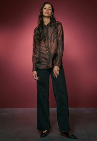 Ruda šilkinė bliuska su tigro raštu, priekyje su mygtukais, ilgomis rankovėmis, derinama su aukštu juosmeniu juodomis plačiomis kelnėmis ir juodais aukštakulniais.