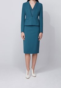 Teal kostymset med en figursydd kavaj med V-hals, kombinerat med en knälång pennkjol. Bärs med vita klackade ankelstövlar.