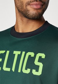 Žalia sportinė marškinė su užrašu "CELTICS" ryškiai laimo žalia spalva. Marškinėliai turi jūrų mėlynos spalvos apvalų kaklą ir tekstūruotą audinį, užtikrinantį kvėpavimą.