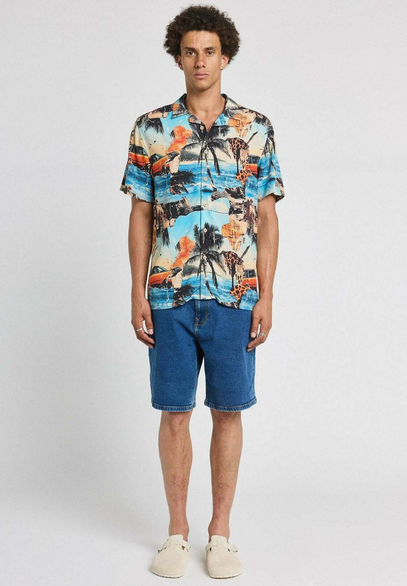 Jeune homme debout portant une chemise à manches courtes à imprimé tropical coloré, un short en jean bleu et des chaussures beige à enfiler sur un fond uni.