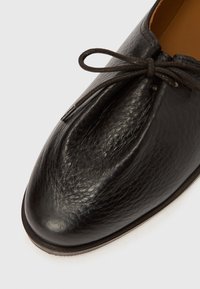 Μαύρο δερμάτινο παπούτσι με στρογγυλεμένη μύτη, υφή φινιρίσματος και καφέ κορδόνια. Διαθέτει μίνιμαλ σχεδιασμό και ανοιχτόχρωμη σόλα.