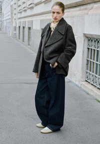 Abrigo de lana gris de gran tamaño, pantalones vaqueros oscuros holgados y zapatillas de crema. Lleva una bufanda ligera alrededor del cuello. Entorno urbano.