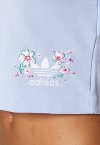 Jasnoniebieska bawełniana tkanina o teksturowanej powierzchni. Zawiera biały haftowany logo Adidas otoczone różowymi i zielonymi wzorami kwiatowymi.