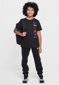 Fekete Nike póló, különböző logónyomtatásokkal, fekete melegítőnadrággal és szürke sportcipővel párosítva. A gyerek egy fekete hátizsákot visz.