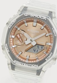 Montre G-SHOCK transparente avec un cadran rose gold, affichage numérique, accents argentés et matériel de protection. Dotée d'un bracelet en résine transparente.