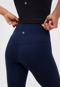 Hænder, der justerer en sort top og marineblå højtaljede leggings med synlige Lululemon-logoer på ryggen ved taljen og den nederste del af ryggen.