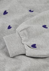 Šedá mikina s manžetou a malými vyšívanými fialovými srdíčky rozptýlenými po látce.