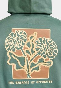 Sudadera verde con un diseño floral texturizado en crema y marrón, con líneas onduladas y un detalle rectangular con borde en la parte posterior.