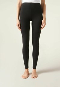 Leggings noirs à côtes avec une taille haute et une coupe cintrée, fabriqués dans un matériau extensible. Pieds nus visibles.
