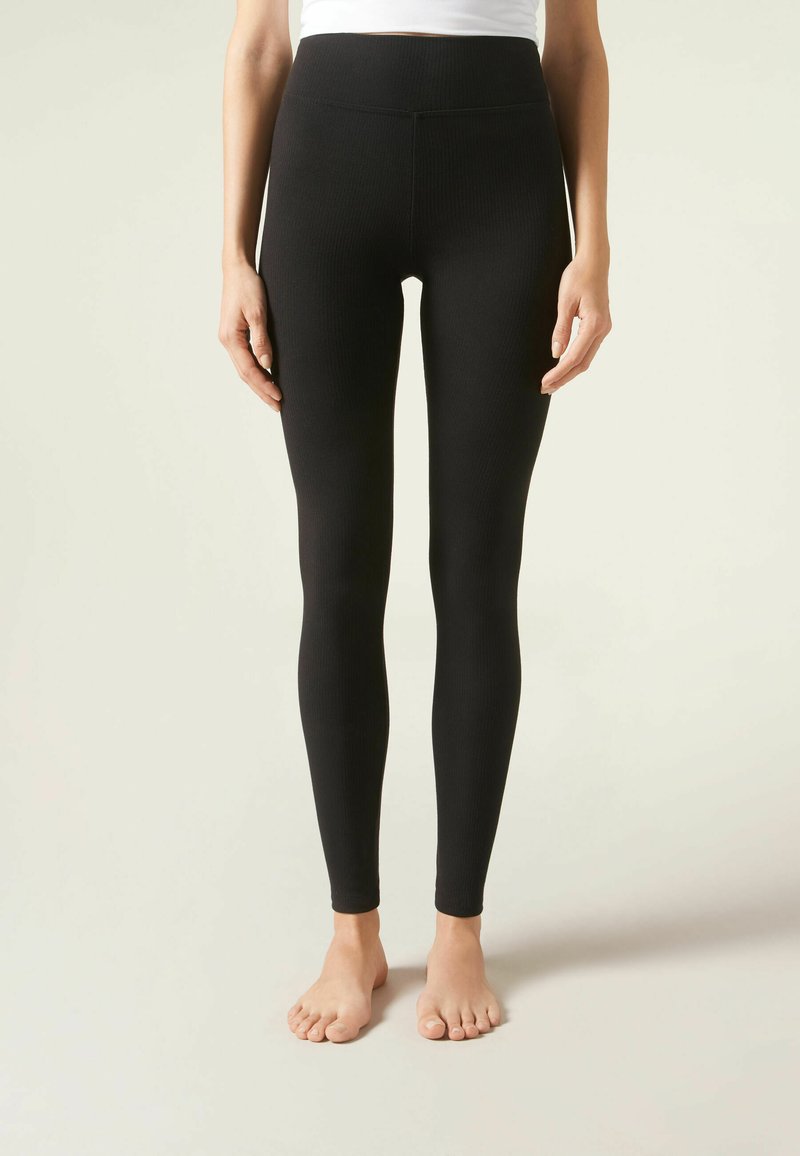 Leggings noirs à côtes avec une taille haute et une coupe cintrée, fabriqués dans un matériau extensible. Pieds nus visibles.