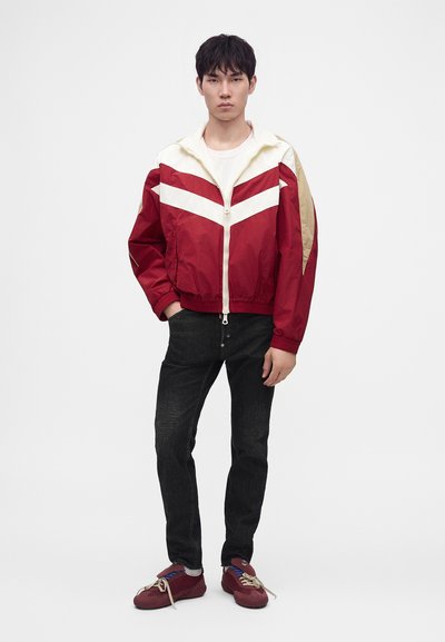 Jeune homme debout portant une veste zippée rouge et blanche, un jean noir et des baskets rouges sur un fond blanc uni.
