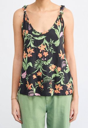 Top de tirantes negro con patrones florales vibrantes en naranja y verde, tirantes ajustables que se atan y un corte holgado, combinado con pantalones verde claro.
