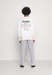 Hvid sweatshirt med grafisk design, der viser en løve og tekst, parret med stribede bukser og sorte sneakers. Bagview på en ensfarvet baggrund.