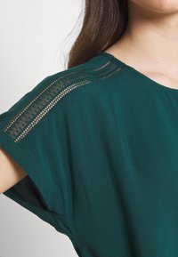 Haut vert à manches courtes avec une coupe décontractée, présentant un détail en dentelle texturée le long de la couture de l'épaule et une texture de tissu lisse.