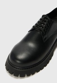 Chaussure en cuir noir avec un bout rond, des coutures détaillées, des lacets plats et une semelle en caoutchouc épaisse et crantée. Fini brillant sur l'ensemble.