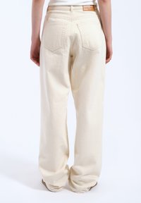 Crème-kleurige wijde jeans van denim, met achterzakken en een tan-label op de tailleband. De textuur lijkt zacht en glad.