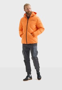 Chaqueta acolchada naranja con capucha, diseño acolchado, cremallera negra y bolsillos, combinada con pantalones cargo grises y zapatillas negras.