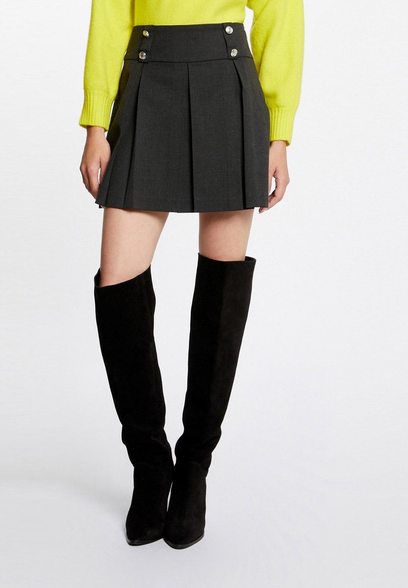 Femme portant une mini-jupe plissée noire avec des boutons, un haut à manches longues jaune et des bottes noires montant jusqu'aux genoux.