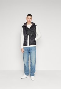 Chaleco acolchado negro con capucha sobre una camiseta de manga larga blanca, combinado con jeans de color azul claro y zapatillas blancas, que lleva un bolso de cintura negro.