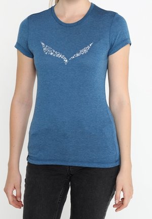 Osoba nosząca dopasowany niebieski t-shirt z białym stylizowanym logo ptaka wykonanym z kwiatowych wzorów na piersi, w połączeniu z czarnymi spodniami.