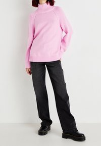 Pull col roulé rose tricoté avec poignets et ourlet côtelés, associé à un jean gris foncé à jambes larges et des chaussures à plateforme noires.