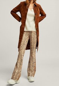 Femme portant un long manteau en daim marron, un pull en tricot crème, un pantalon à imprimé serpent et des baskets chunky blanches, se tenant les mains sur les hanches.