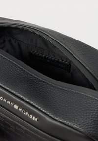 Mustapintainen nahkainen pussi, jossa on avoin vetoketju, joka paljastaa kangasvuoren. Edessä ja sisäisessä tarrassa on Tommy Hilfiger -logo.