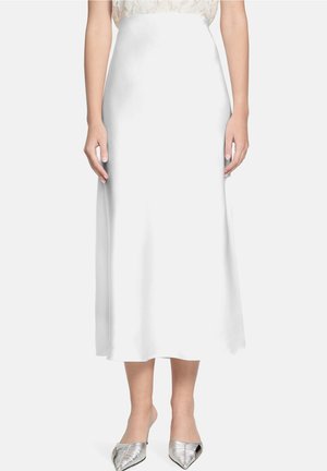 Vrouw draagt een witte satijnen midi-rok met hoge taille, een kanten top en zilveren puntige instapschoenen, staand tegen een witte achtergrond.