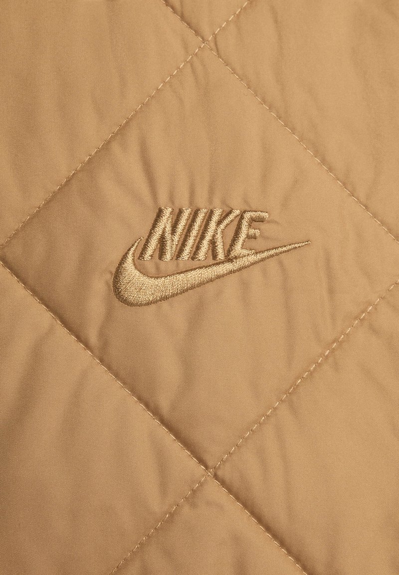 Beżowy materiał pikowany z haftowanym złotym logo Nike, z diamentowym wzorem i gładką teksturą.