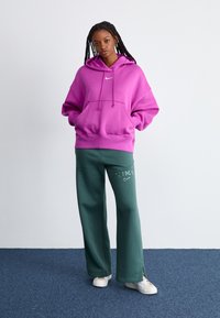 Sudadera oversized morada con un bolsillo tipo canguro en la parte delantera y un pequeño logo blanco, combinada con pantalones de chándal verde de pierna ancha con letras blancas.