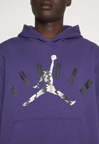 Personne portant un sweat à capuche violet avec le texte noir "JORDAN" et un logo Jumpman camouflage noir et blanc sur la poitrine.