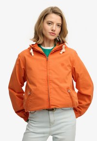 Giacca arancione con zip e cappuccio, con coulisse bianche regolabili, tasche frontali e polsini elasticizzati. Indossata sopra una maglia verde.