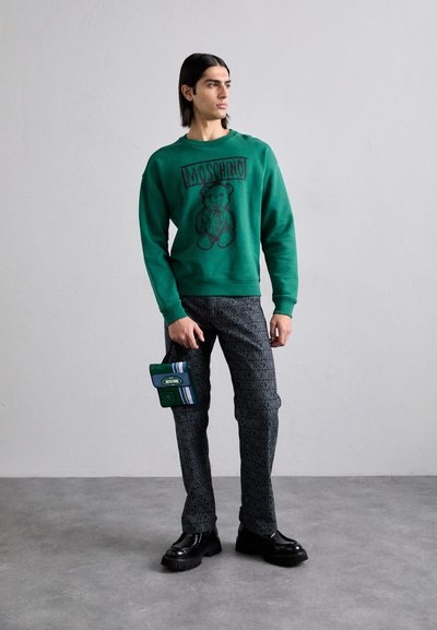 Un hanorac verde cu un grafic cu un urs, asociat cu pantaloni negri cu un model care prezintă textul "Moschino". Accesoriile includ o gentuță verde cu dungi.
