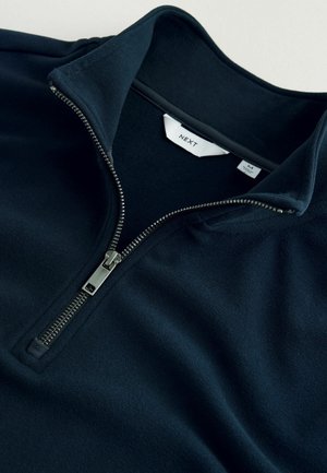 Marineblå fleece-pullover med høj krave, halv lynlås og glat tekstur. Har en sølvfarvet lynlås og mærketag indeni.